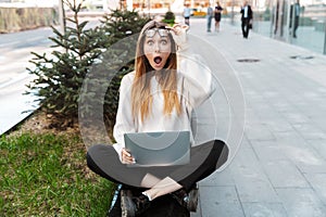 Shocked young business woman posing sitting outdoors near business center wearing eyeglasses using laptop computer