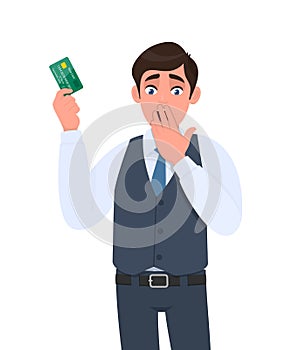 Shocked young businessman showing credit card and covering mouth with hand. Frustrated person in waistcoat holding debit, ATM card