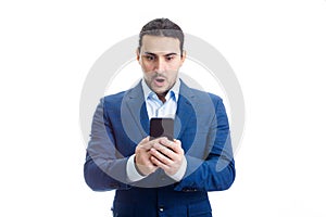 Shocked young businessman using mobile phone staring with big eyes and mouth open isolated on white wall background, with copy