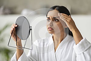 Shocked young eastern woman looking at mirror, touching her face