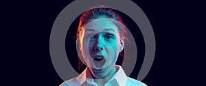 Shocked young girl in white shirt looking at camera with open mouth isolated on dark background in blue neon light