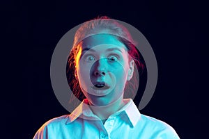 Shocked young girl in white shirt looking at camera with open mouth isolated on dark background in blue neon light