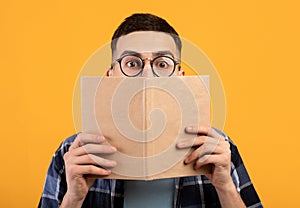 Shocked young guy in glasses hiding behind book, having problem studying for college exams on orange background