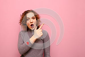 Shocked young lady points her finger at copy space and looks into camera with emotional face. Surprised curly lady stands on a