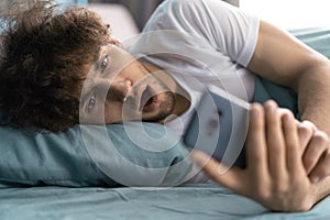 Shocked young man in bed looking at smartphone screen at home, overslept. Guy is late for appointment or work or class