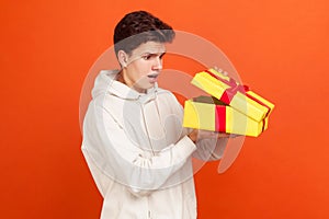 Shocked young man in casual white sweatshirt with hood glancing inside present box, disappointed with gift, unjustified