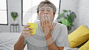 Shocked young man, coffee in hand, sitting quietly on bedroom bed, covering mouth in surprised fear, regret apparent in his eyes