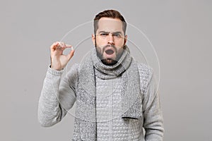 Shocked young man in gray sweater, scarf isolated on grey wall background. Healthy lifestyle ill sick disease treatment