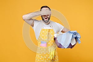 Shocked young man househusband in apron hold basket with clean clothes while doing housework isolated on yellow