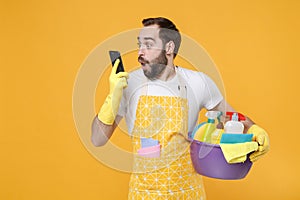 Shocked young man househusband in apron rubber gloves hold basin with detergent bottles washing cleansers doing