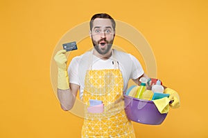 Shocked young man househusband in apron rubber gloves hold basin with detergent bottles washing cleansers doing