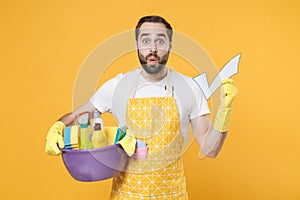 Shocked young man househusband in apron rubber gloves hold basin detergent bottles washing cleansers doing housework