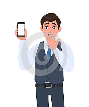 Shocked young man in vest suit showing mobile, cell or smartphone. Frustrated trendy person covering hand on mouth. Male character