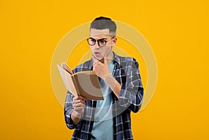 Shocked young student in glasses reading textbook, having difficulty studying for exam on orange studio background