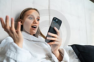 Shocked young woman in bathrobe looking at mobile phone in bed at home