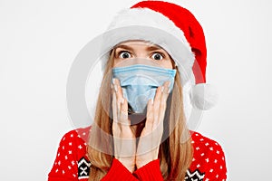 Shocked young woman wearing a Santa Claus hat and a medical protective mask on her face, on a white background, Celebrating New