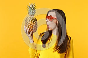 Shocked young woman in funny glasses looking on fresh ripe pineapple fruit in hand isolated on yellow orange background