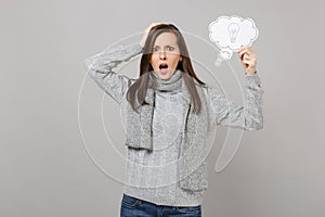 Shocked young woman in gray sweater, scarf put hand on head, holding say cloud with lightbulb isolated on grey wall