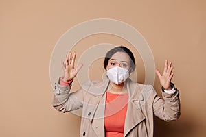 Shocked young woman in medical mask, gesturing, expressing stupefaction looking at camera, on isolated beige background