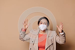 Shocked young woman in medical mask, gesturing, expressing stupefaction looking at camera, on isolated beige background photo