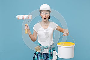 Shocked young woman in protective helmet hardhat hold paint bucket, roller for wall painting isolated on blue background