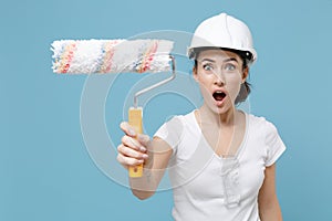 Shocked young woman in protective helmet hardhat hold paint roller for wall painting isolated on blue background in