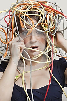 Shocked young woman`s head tangled in colorful cables over gray background