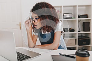 Shocked young woman in striped shirt looking at laptop screen and holding glasses. Curly black girl sitting at her