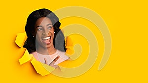 Shocking happy laughing millennial black lady with open mouth looks through hole in yellow paper