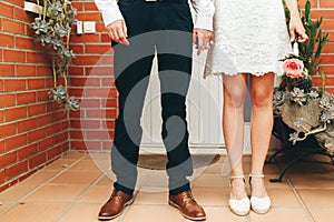 Shoes of groom and bride and her small wedding bouquet of pink roses with clasped hands after the ceremony. Wedding concept.