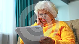 shoked amazed old woman white gray-haired sitting on the sofa in living room with bills