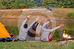 Shoot from back. A group of happy friends camping at riverside, dancing hold hands up  and enjoy view. Holidays fun