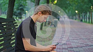shooting advertising, a young guy in the park sits on a bench in black clothes and plays on the phone.