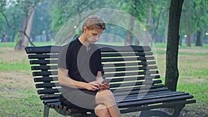 shooting advertising, a young guy in the park sits on a bench in black clothes and plays on the phone.