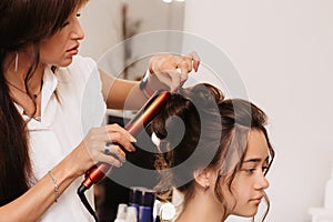 Shooting in a beauty salon. A hair stylist makes a hairstyle for a young dark-haired girl with the help of a hair styler