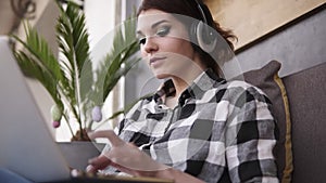 Shooting from below of a charming brunette sitting on a sofa. Girl in headphones typing on laptop, smiling