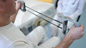 Shooting over shoulder of motivated man exercising with resistance band and blurred unrecognizable doctor at background