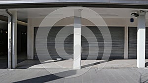 Shop with shutters lowered, under a porch with columns. White wall, shadows and in front a tiled pavement.