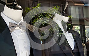 Shop window of mens tailor shop showing 2 mannequins in tuxedos.