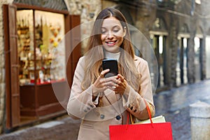 Shopper woman buying online with smart phone in old city street in winter