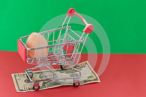 A shopping basket on a red and green background with a plastic egg stands on a banknote. The concept of unnatural products with