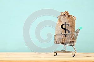 shopping cart with bag full of money with dollar sign over blue wooden background.
