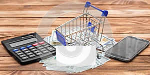 Shopping cart with blank business card, money, calculator and smartphone on the table