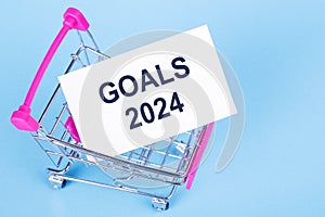 Shopping cart on blue background with paper card inside. The inscription on the card GOALS 2024. Concept of goals for the next