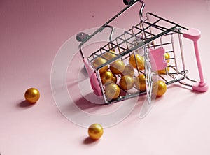 Shopping cart. Fallen Supermarket trolley full of golden balls on pink background. Consumerism concept photo.