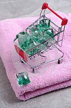 Shopping cart filled with laundry capsules stands on pink, terry towel.