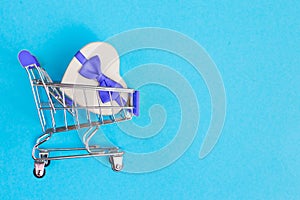 Shopping cart with gift box on blue background. Shopper accessory, consumerism and commerce symbol