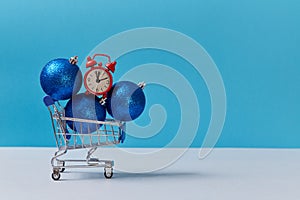 Shopping cart with glitter balls and red clock. Twelve oclock. New year and xmas. Copy space. Isolated on blue backdrop