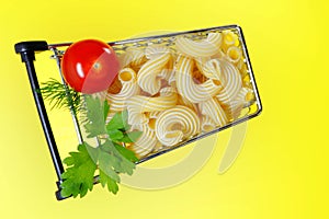 A shopping cart with Italian pasta, ripe cherry tomatoes, parsley and dill. Bright yellow background. Copy space.