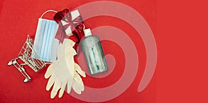 Shopping cart with medical mask and gel, gloves and gift box on red background. Christmas and coronavirus  concept